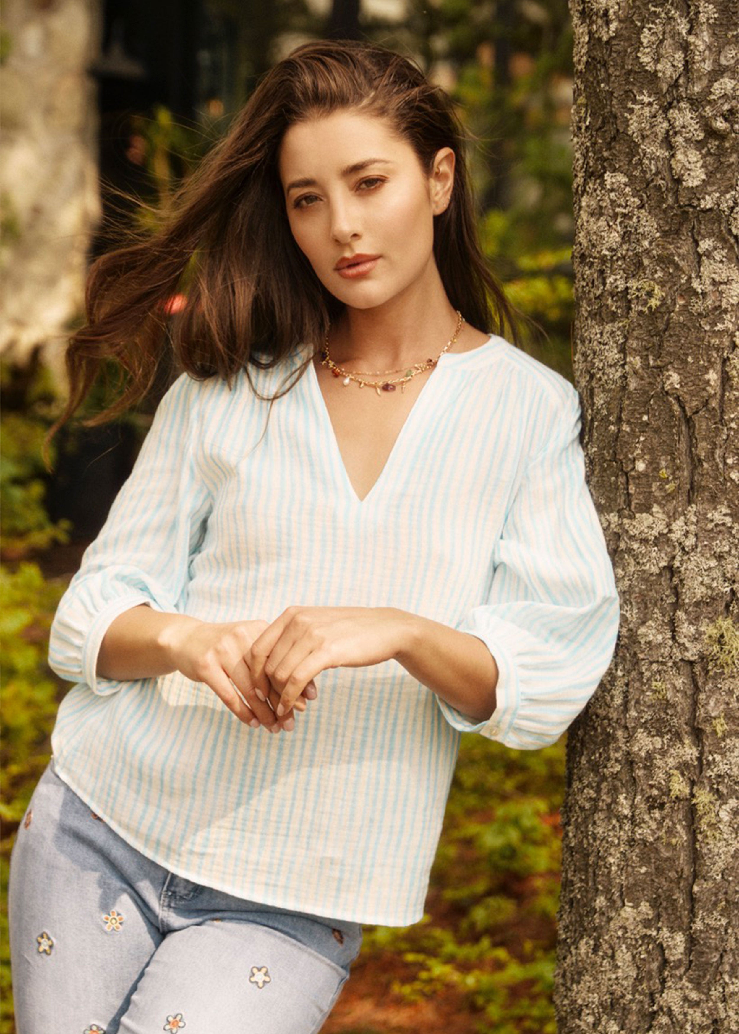Model leaning against tree in light blue striped blouse, showcasing casual elegance by French Dressing Jeans (FDJ)
